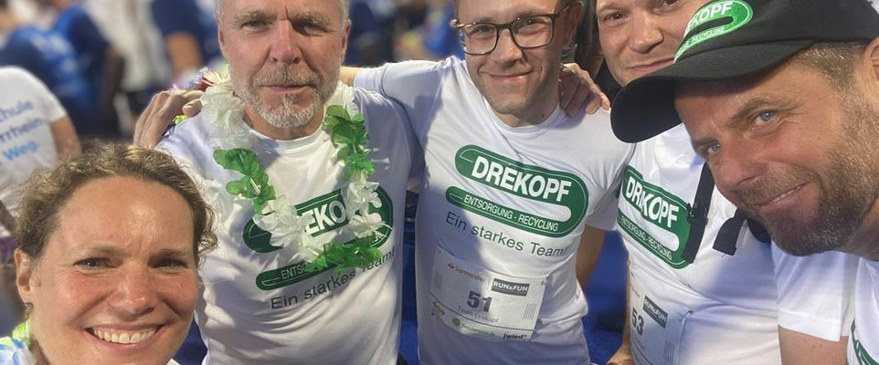 Fünf Personen, vier Männer und eine Frau, posieren eng beieinander und lächeln für ein Gruppenfoto in einem Stadion. Alle tragen weiße Hemden mit grünem Drekopf-Logo und vertreten stolz das Team Drekopf beim Run & Fun Lauf 2023.