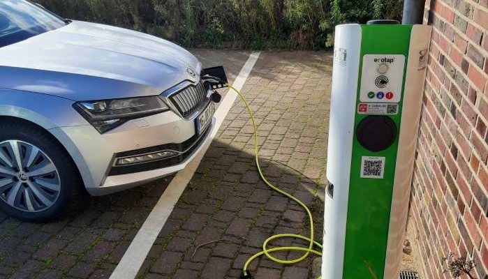 Ein silbernes Auto wird auf einem markierten Platz geparkt und mit einem gelben Kabel an eine Ladestation für Elektrofahrzeuge angeschlossen, die Co2-Reduktion unterstützt. Die grün-weiße Station ist mit sichtbaren Tasten und einem QR-Code versehen.